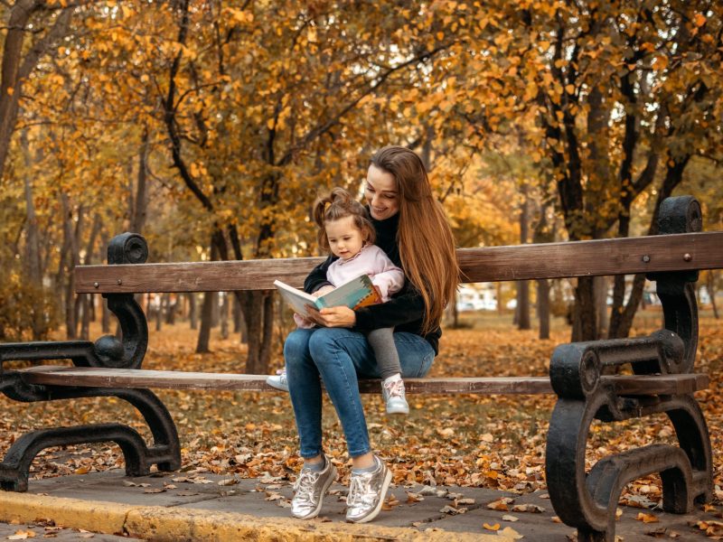 Young female woman babysitter and toddler baby girl read book in autumn park. Happy family mom and toddler outdoors in fall park.